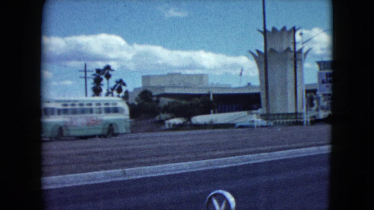 1959 Nevada 8mm frame showing a roadside view with a classic bus, palm trees, wide street, parked cars, and a tall modernist tower.