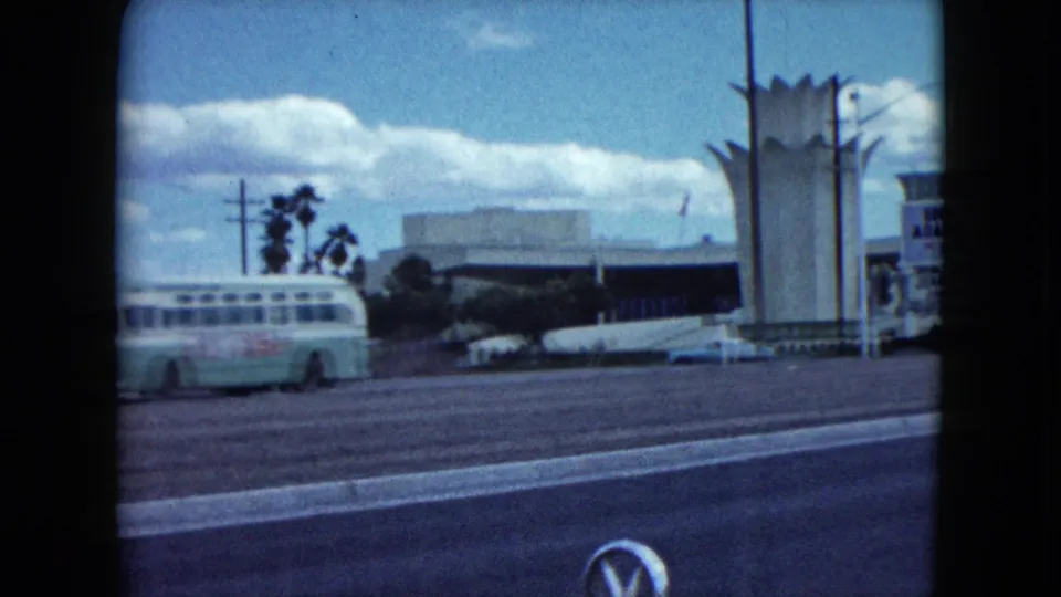 1959 Nevada 8mm frame showing a roadside view with a classic bus, palm trees, wide street, parked cars, and a tall modernist tower.