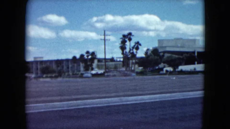 1959 Nevada roadside frame with a broad paved street, palm trees, parked cars, low commercial buildings, and utility poles.