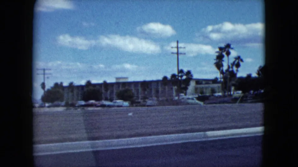 1959 Nevada home-movie view of a town street with low-rise buildings, palm trees, parked cars, utility poles, and open roadway.