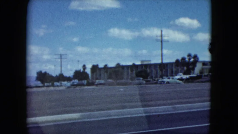 1959 Nevada 8mm frame showing a moving-car view across a wide street toward low buildings, parked cars, palms, and utility poles.