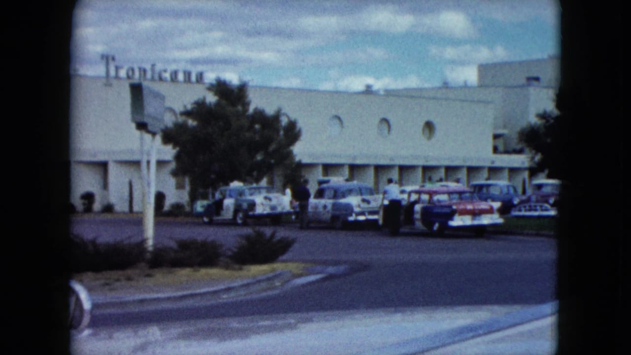 Pale mid-century building with Tropicana sign, parked classic cars, trees, curb, and cloudy sky.