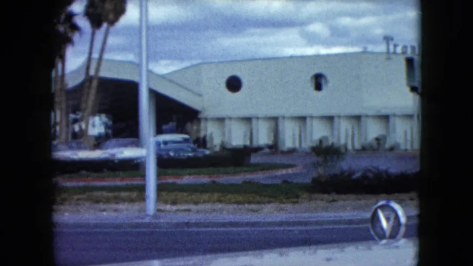 1959 Nevada frame of a mid-century roadside building with round windows, palm trees, parked cars, mountains, and a street foreground.