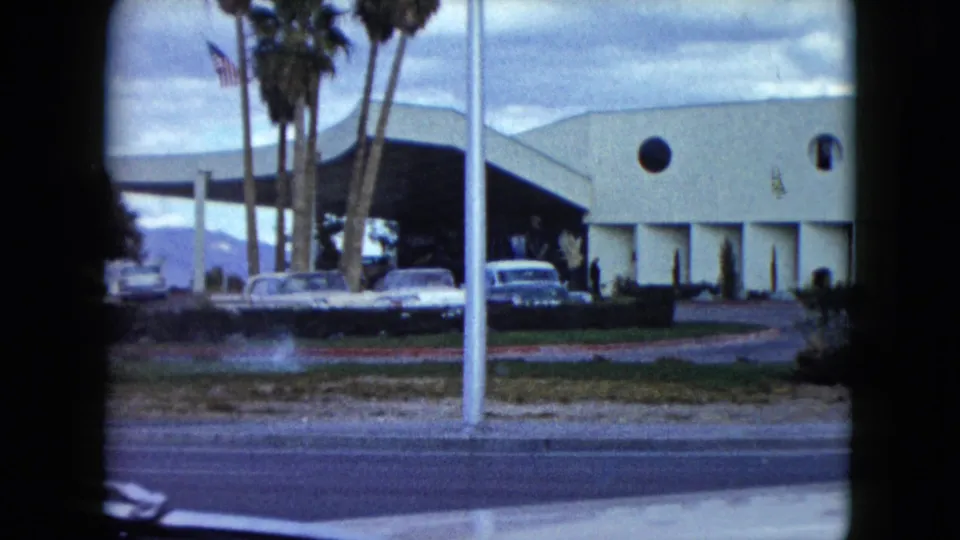 1959 Nevada home-movie frame showing a pale building with a round window, palm trees, parked cars, a flagpole, and distant mountains.