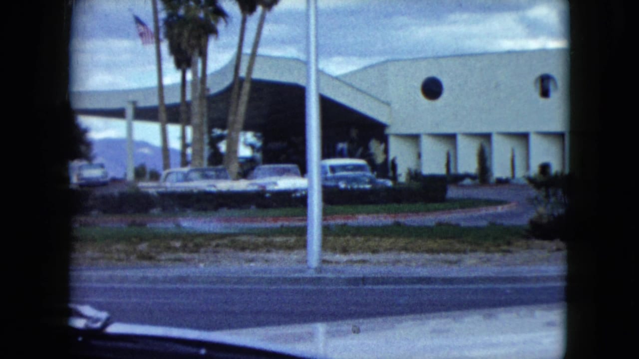 1959 Nevada road view of a mid-century style building, palm trees, a flagpole, and parked period cars seen from inside a vehicle.