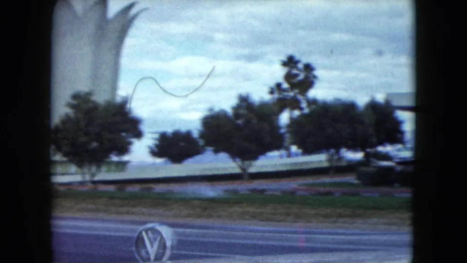 White lotus-like monument in Nevada, filmed across a road with trees, lawn, and a partial parked car in a soft 8mm frame.