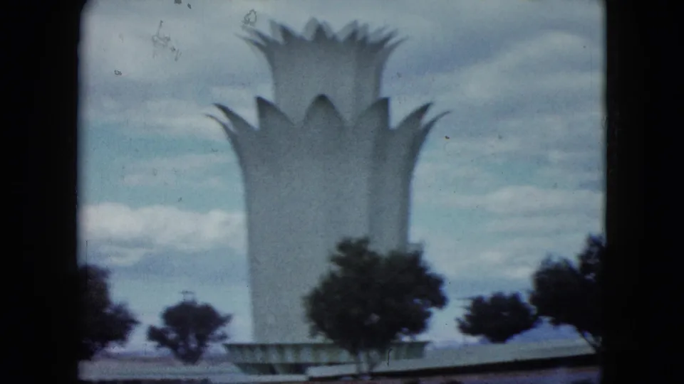 Closer 1959 home-movie view of a white lotus-like monument rising behind roadside trees under a cloudy sky in Nevada.