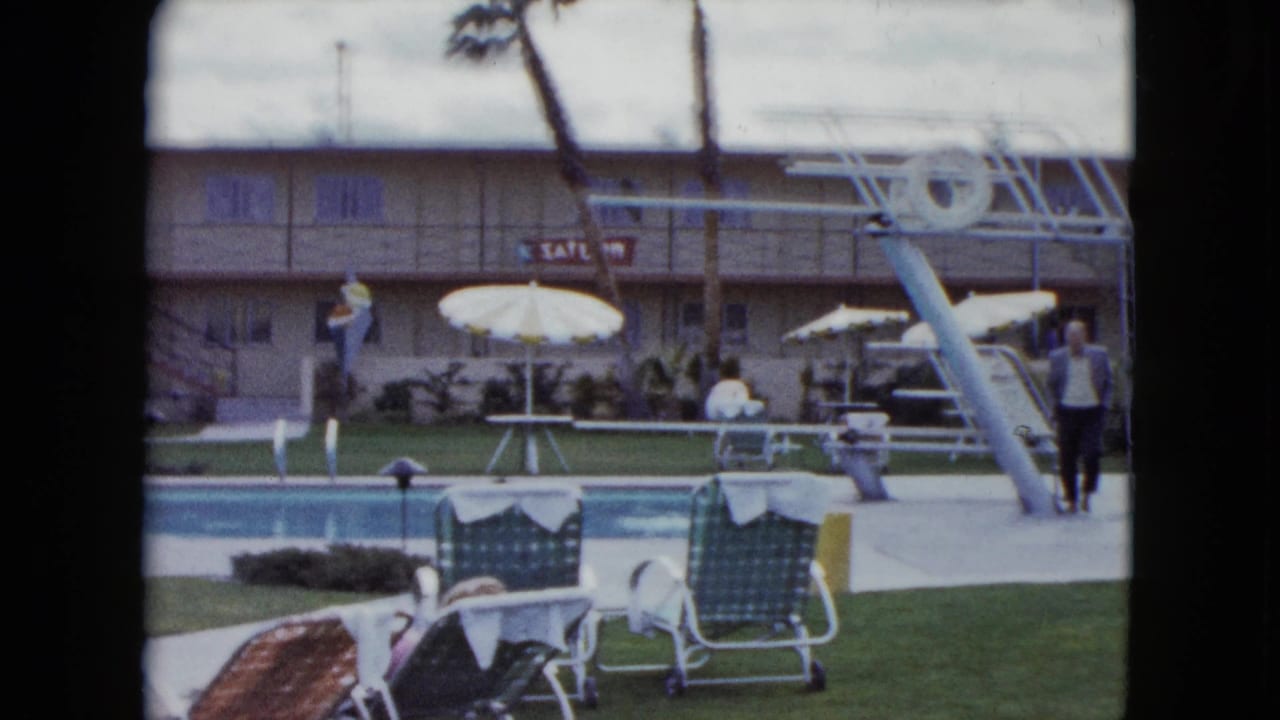 1959 Nevada home-movie frame showing a motel swimming pool with white umbrellas, lounge chairs, a diving board, palm trees, and visible guests.