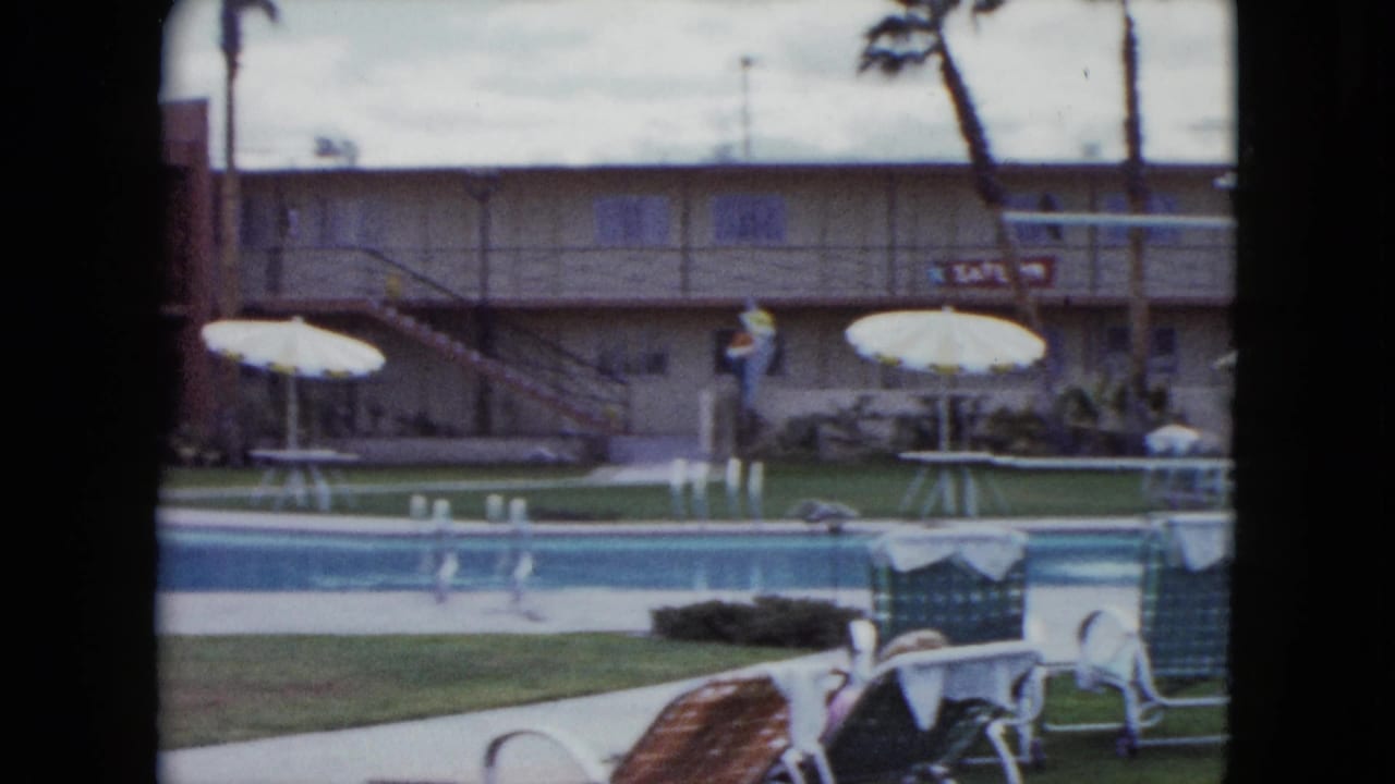 Swimming pool and lawn with white umbrellas, lounge chairs, palm trees, and a motel building behind them.