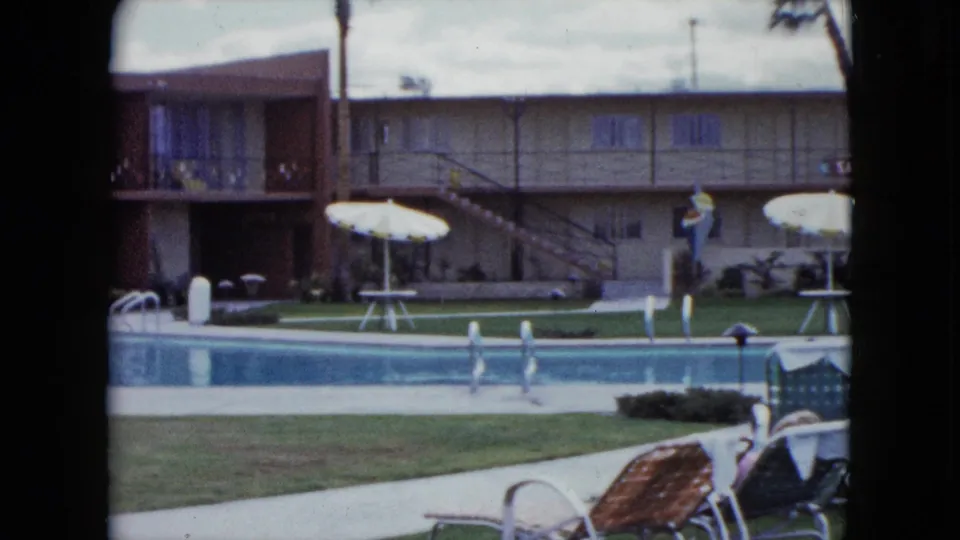 1959 Nevada home-movie view of a motel pool courtyard with white umbrellas, exterior stairs, lounge chairs, lawn, and calm blue water.