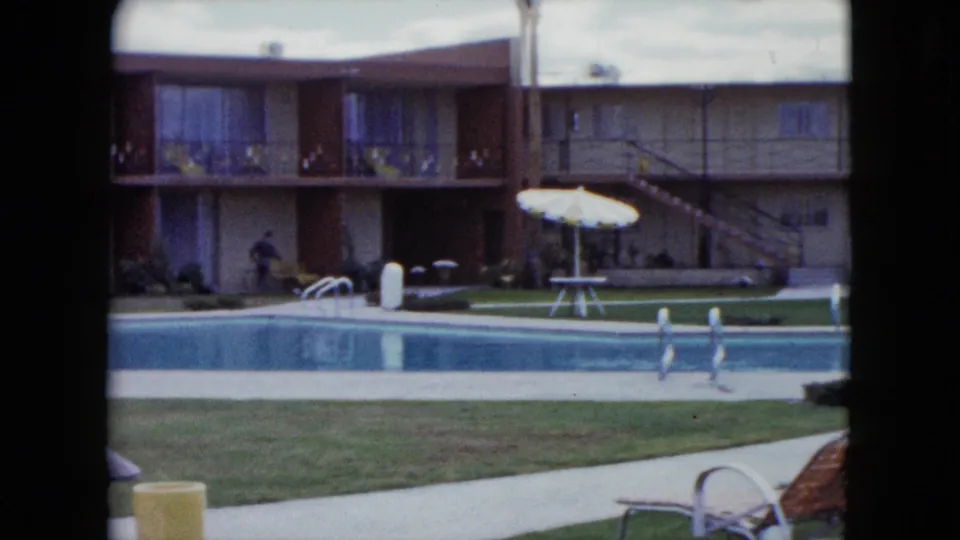 1959 Nevada frame showing a broad motel pool area with white umbrellas, balconies, exterior stairs, lawn, and vintage lounge seating.