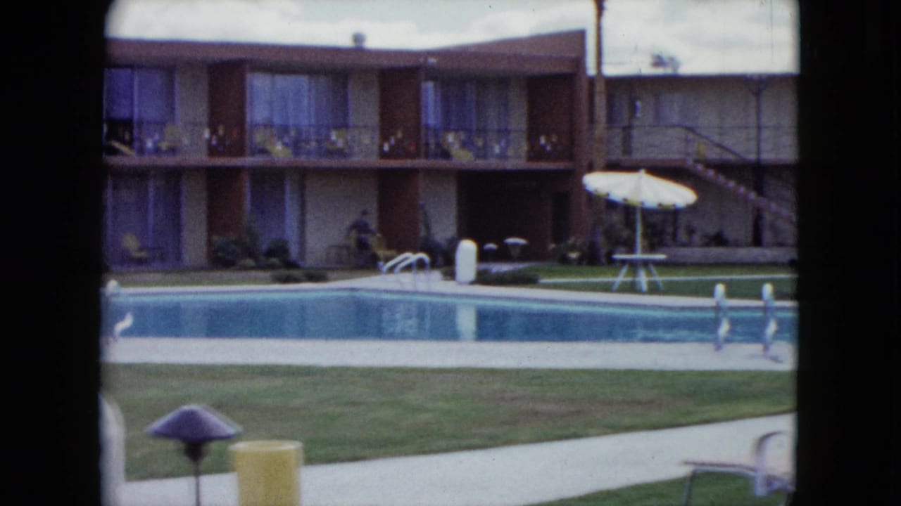 1959 Nevada home-movie frame of a quiet pool courtyard with a two-story motel or apartment building, lawn, and patio umbrella.