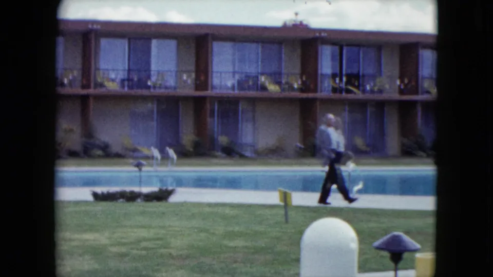 In a 1959 Nevada home-movie frame, a man walks past the pool as balcony rooms, lawn, garden lights, and outdoor furniture remain in view.