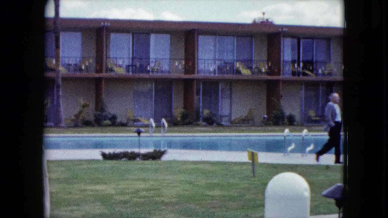 1959 Nevada home-movie frame showing a motel pool courtyard with lawn, balcony rooms, pool ladders, and adults moving through the scene.
