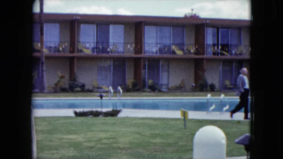 1959 Nevada home-movie frame showing a motel pool courtyard with lawn, balcony rooms, pool ladders, and adults moving through the scene.