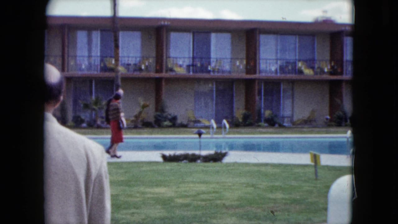 1959 Nevada poolside motel scene with a foreground man in a light jacket and a woman in a red skirt walking along the courtyard.