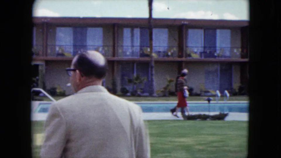 1959 Nevada 8mm frame with a bald man in glasses in the foreground and a woman walking beside a motel swimming pool.