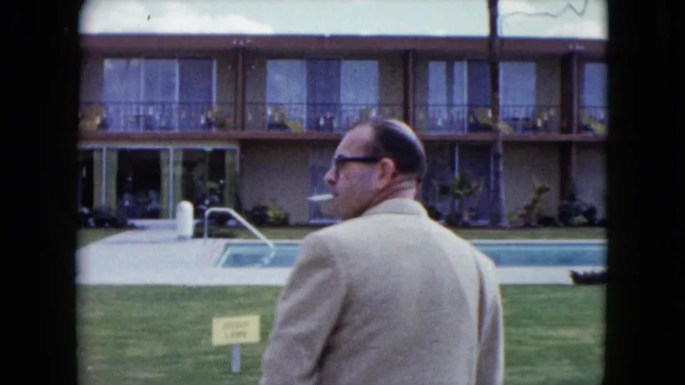 1959 Nevada home-movie frame showing a man in a light suit and glasses smoking beside a mid-century motel pool courtyard.