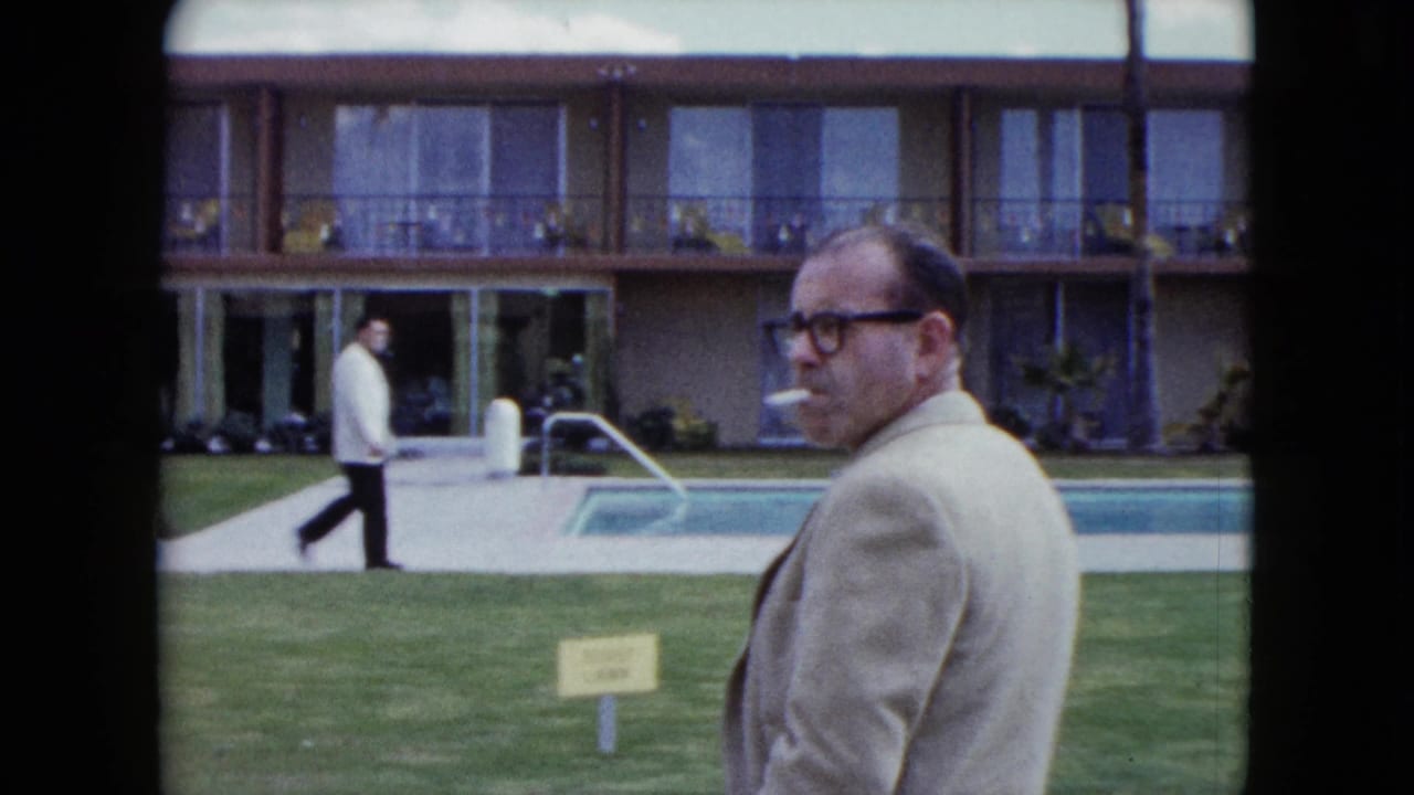 In 1959 Nevada home-movie footage, a man in a light suit smokes near a motel pool while another guest walks past the two-story rooms.