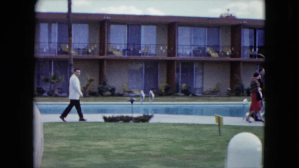 A 1959 Nevada home-movie frame shows a man walking along a motel pool, with balcony rooms, lawn signs, and additional guests near the edge.