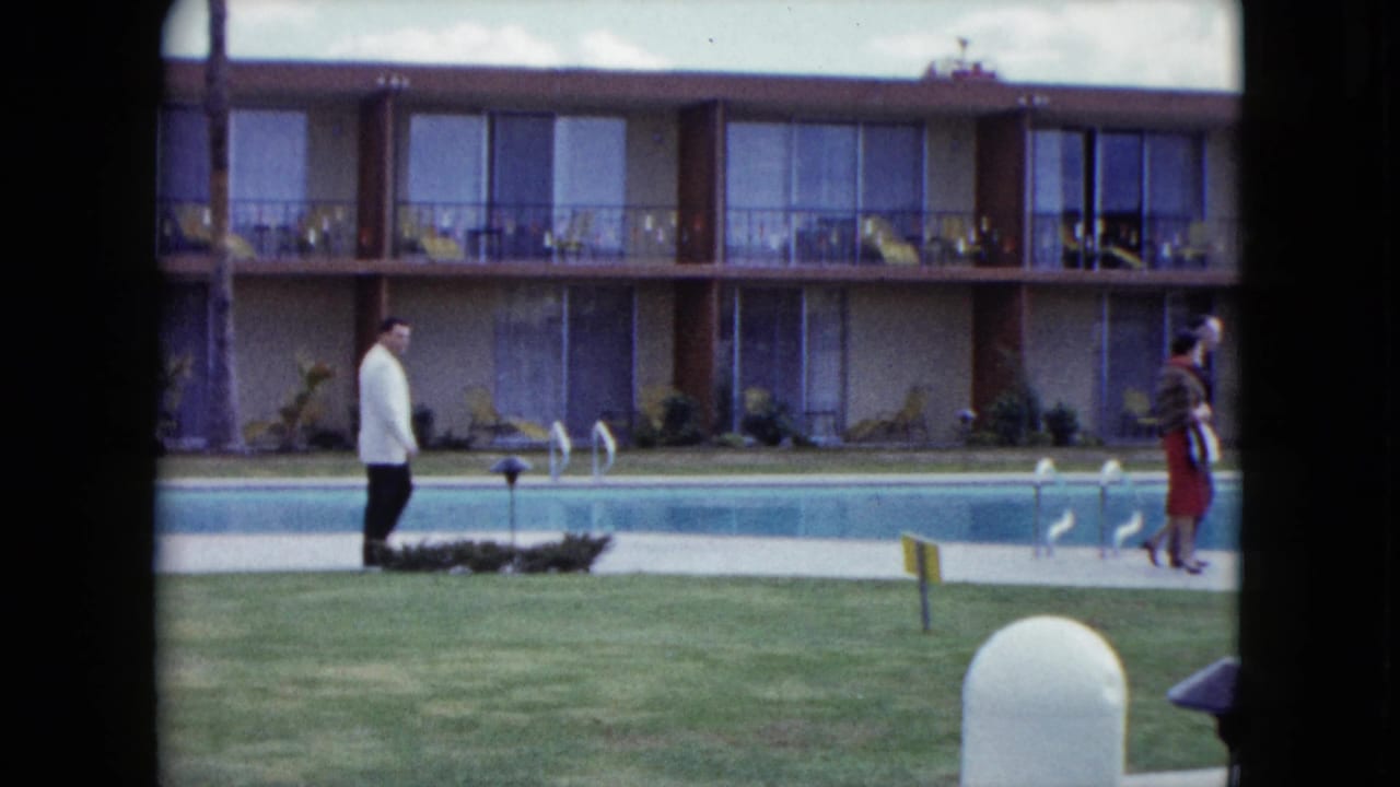 Man in a white jacket walks beside a motel swimming pool as a woman passes nearby.