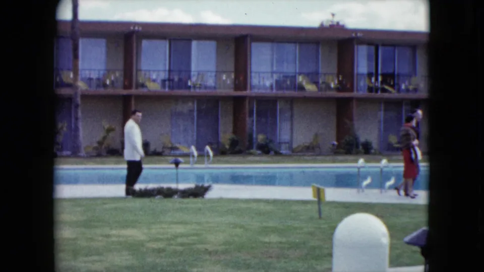 In 1959 Nevada home-movie footage, a man in a white jacket walks along a motel pool while a woman in a red skirt moves across the courtyard.