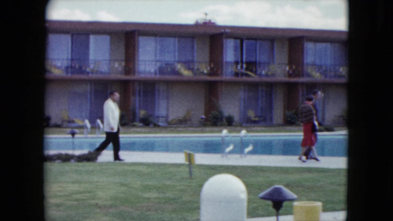Man in a white jacket and woman in a red skirt walk along a motel pool deck.