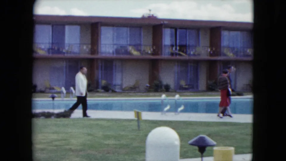 A man in a white jacket and a woman in a red skirt move past a swimming pool at a two-story Nevada motel courtyard in 1959 footage.