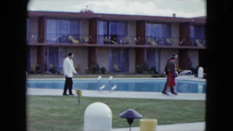 Two adults walk beside a motel swimming pool, with two-story balconies, lawn, pool lights, and mid-century courtyard details visible.