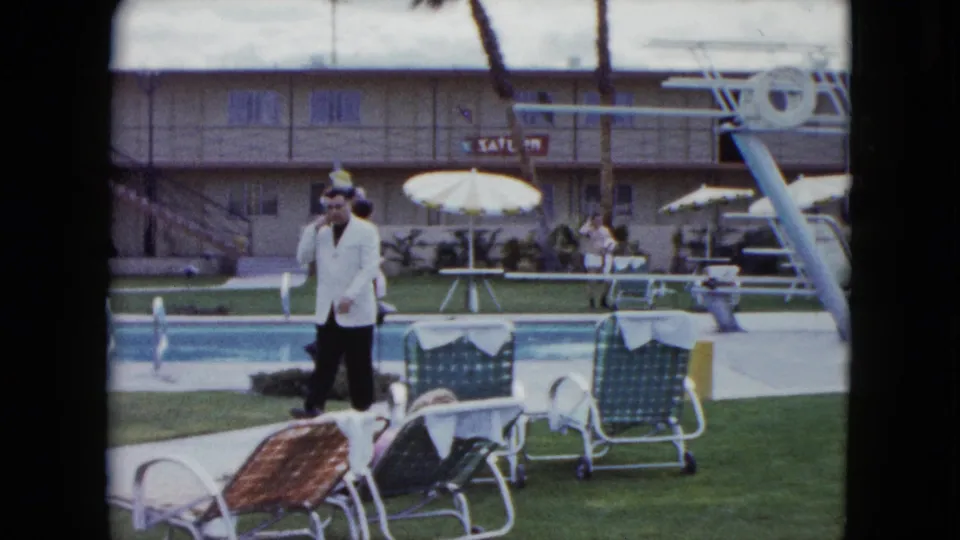 A man in a white jacket stands by lounge chairs near a motel swimming pool, with umbrellas, a diving board, and courtyard buildings visible.
