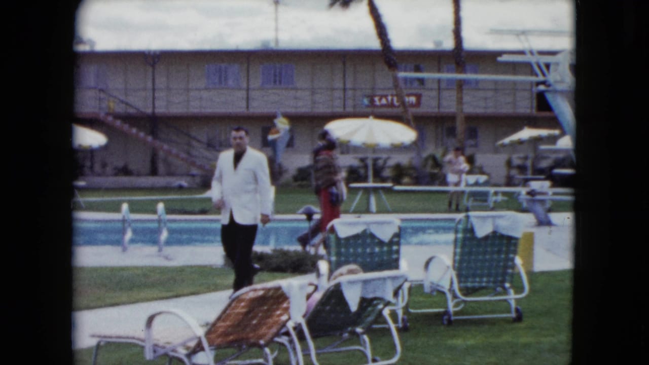 Man in a white jacket walks beside a motel swimming pool with lounge chairs and umbrellas.