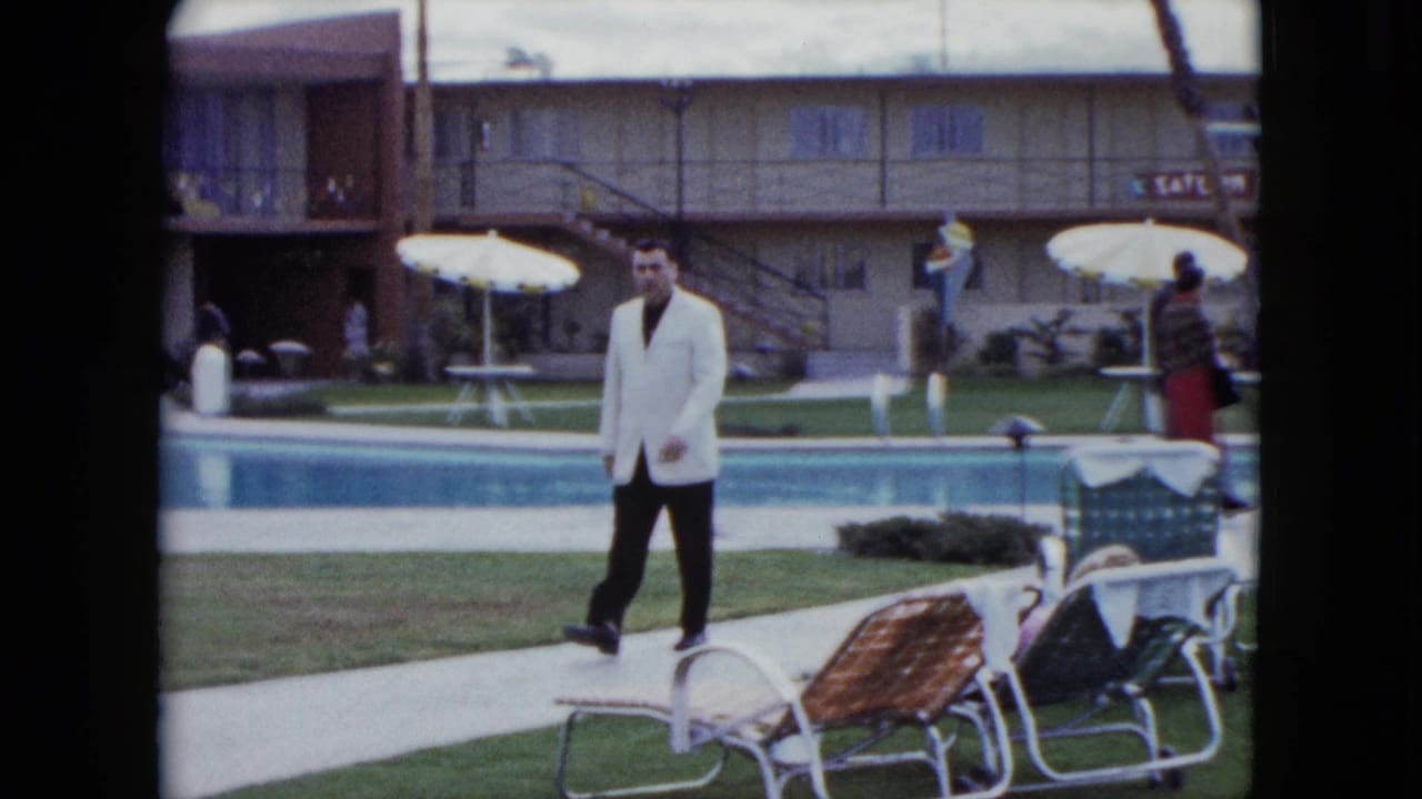 The man continues along the poolside path, with motel balconies, umbrellas, chairs, and guests behind him.