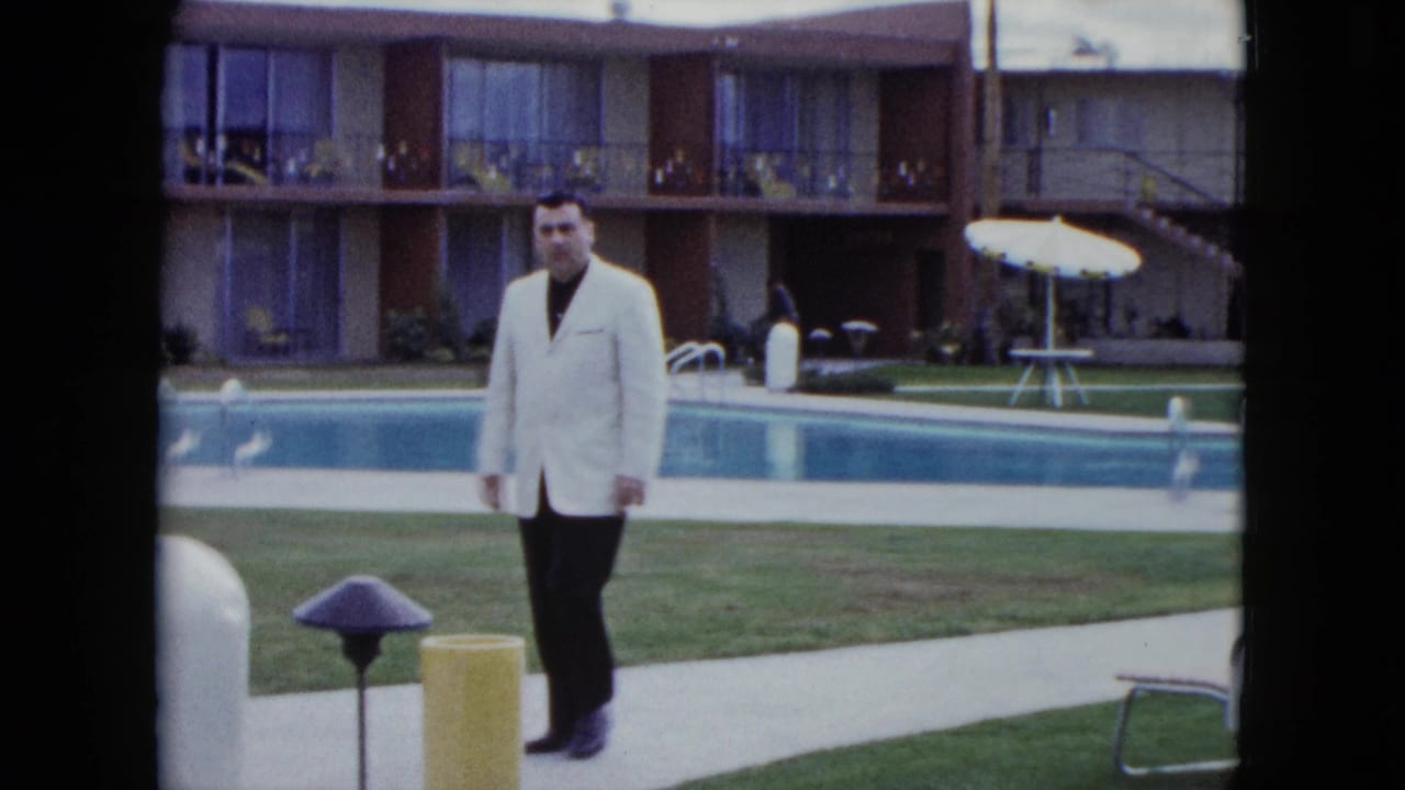 Man in a light jacket walking beside a motel swimming pool and lawn.