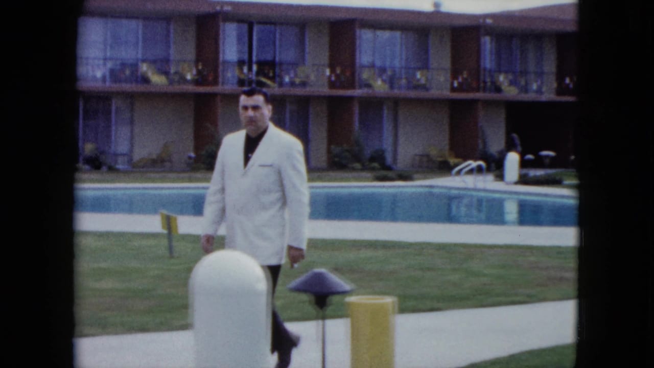 Man in a light jacket walking near poolside fixtures in a motel courtyard.