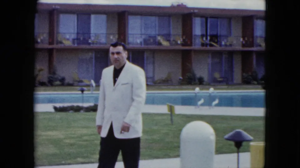 A man in a light jacket stands or slows near the motel pool, with lawn, pool edge, balcony rooms, and courtyard furnishings in view.