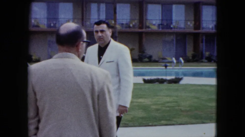 Two adult men appear to meet or speak near the motel swimming pool, with the two-story balcony building and lawn in the background.
