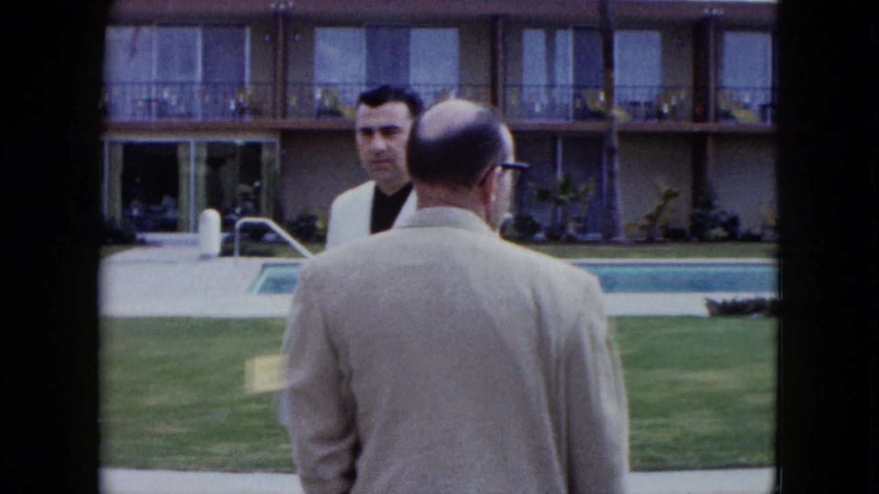 Two men near a resort pool, one facing away and one in a white jacket facing the camera.