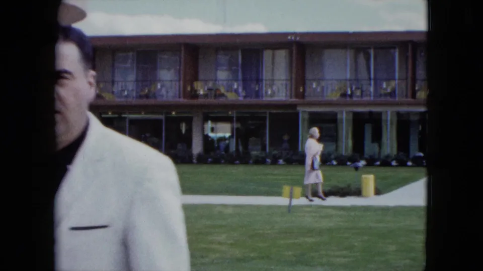 1959 Nevada poolside home-movie frame with a cropped man in white and a woman walking near resort balconies.
