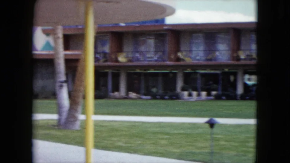 1959 Nevada resort view showing lawn, pool edge, walkway, palm trunk, and balcony rooms in soft 8mm color.