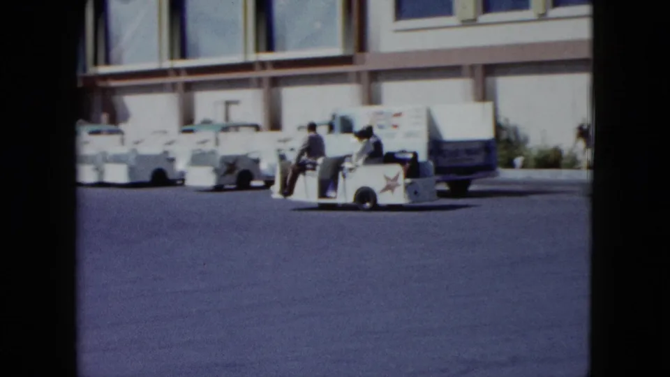 The white resort shuttle cart continues along the paved drive, with riders, parked cars, and building windows visible behind it.