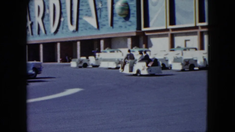 A white shuttle cart with seated riders moves across the resort drive beneath large partial signage and parked cars.