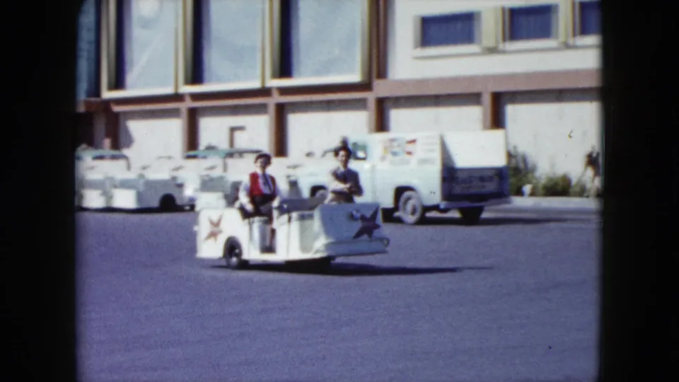 Two people ride a white star-marked cart across the Stardust Hotel lot, with parked cars and a light-colored service vehicle behind them.