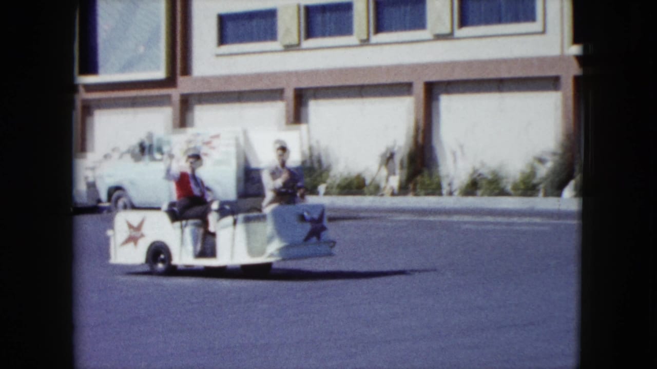 In Nevada 1959 footage, two men ride a small white trolley past a modern building while one turns toward the camera.