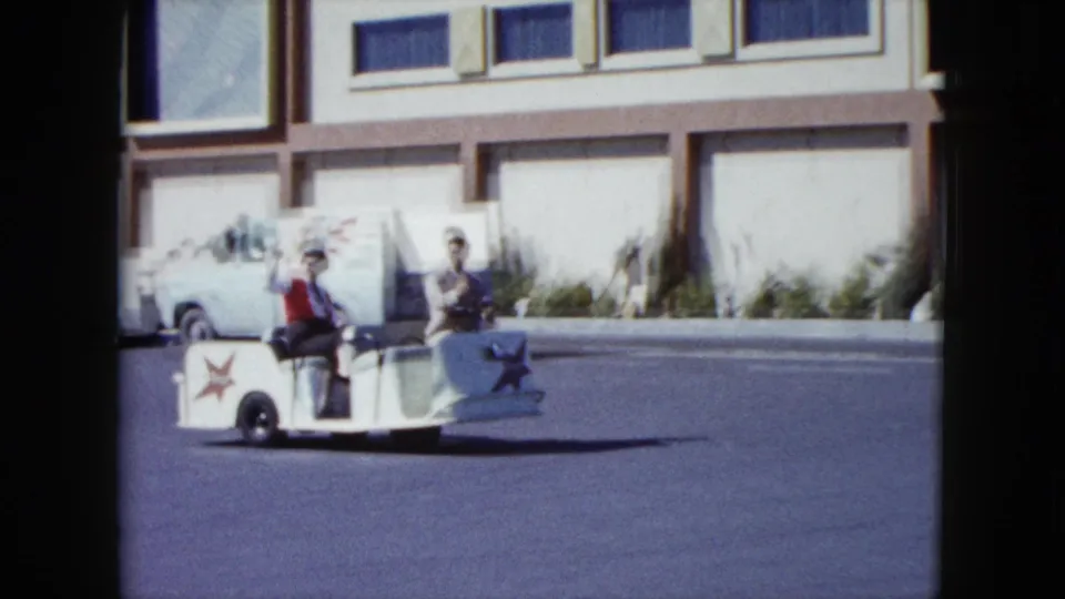 In Nevada 1959 footage, two men ride a small white trolley past a modern building while one turns toward the camera.