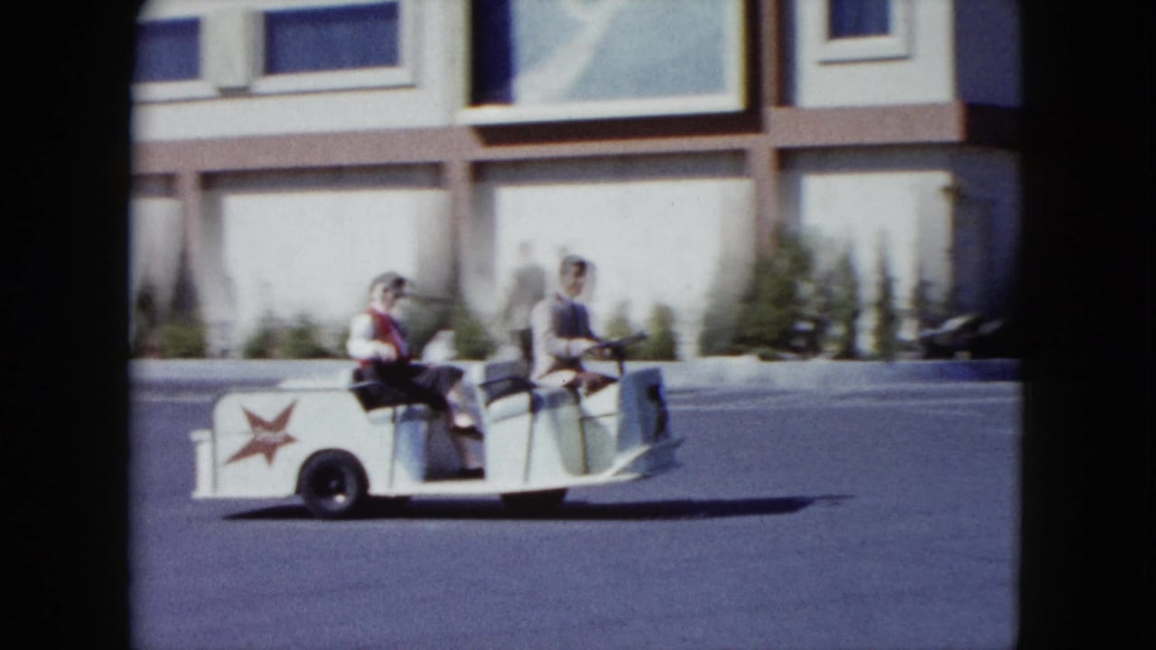Two adult men continue past the camera in a small white trolley with red star logos in Nevada 1959 home-movie footage.