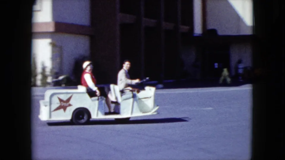 A passenger in a red vest and a driver in a light shirt ride past a shadowed entrance in 1959 Nevada footage.