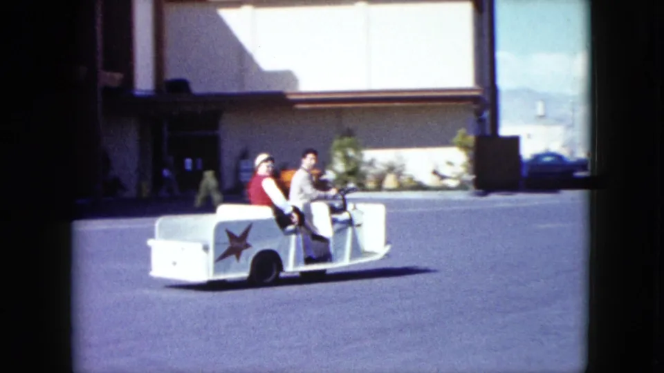 The small white trolley moves farther across the Nevada parking lot, with two men seated and red star markings visible.