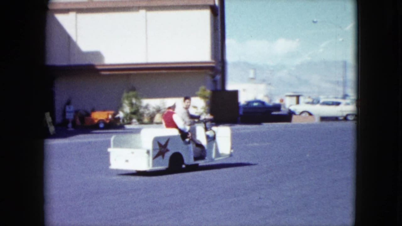 In 1959 Nevada footage, a man and woman ride a small white guide cart past a building edge, with parked cars and mountains behind them.