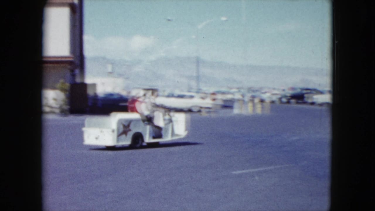 The guide cart continues across a Nevada parking lot, with two adults aboard and rows of mid-century cars under a bright sky.