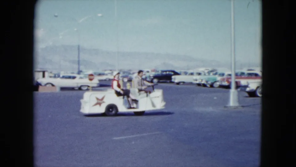 A man and woman ride a white star-marked guide cart through a Nevada lot, with many 1950s automobiles and a light pole visible.